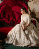 Mahira khan 3 Pcs Ivory ensemble features an Organza ivory shoulder cut-out pishwas with an attached raw silk underlayer, showcasing embroidery on the blouse and detailed borders at the hem. The shirt has fitted three-quarter sleeves and a halter neckline on the upper layer. It transitions into heavily embroidered raw silk gharara pants. Complemented by an Organza ivory dupatta with spray details, a four-sided border, and different shades adding depth and contrast