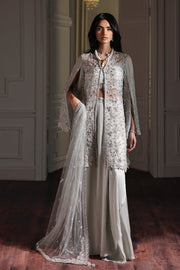Woman wearing a traditional outfit with a long embroidered jacket and white pants in an elegant room.