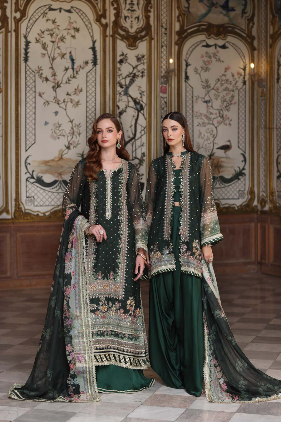 Two women in traditional green and black embroidered outfits standing in an ornate room.