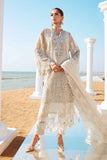 A woman posing in a white and gold embroidered lawn three-piece suit with organza details, including a shirt, trousers, and a dupatta, against an outdoor backdrop with columns and a clear sky.