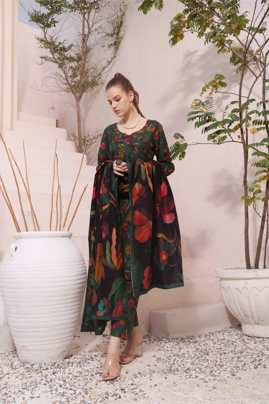 Woman in a long, dark green dress with colorful floral pattern standing in an outdoor setting with plants and white vases.