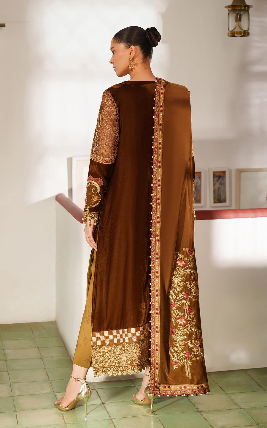 Woman wearing a brown and gold traditional outfit with intricate patterns in a minimal indoor setting.