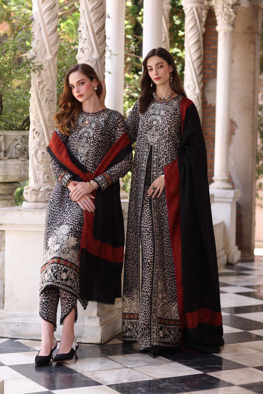 Two women in traditional outfits standing in a decorative setting with columns and greenery.