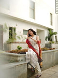 Yashma Gill sitting on a ledge with a phone to her ear, wearing a white outfit and red shawl, in front of a modern building.