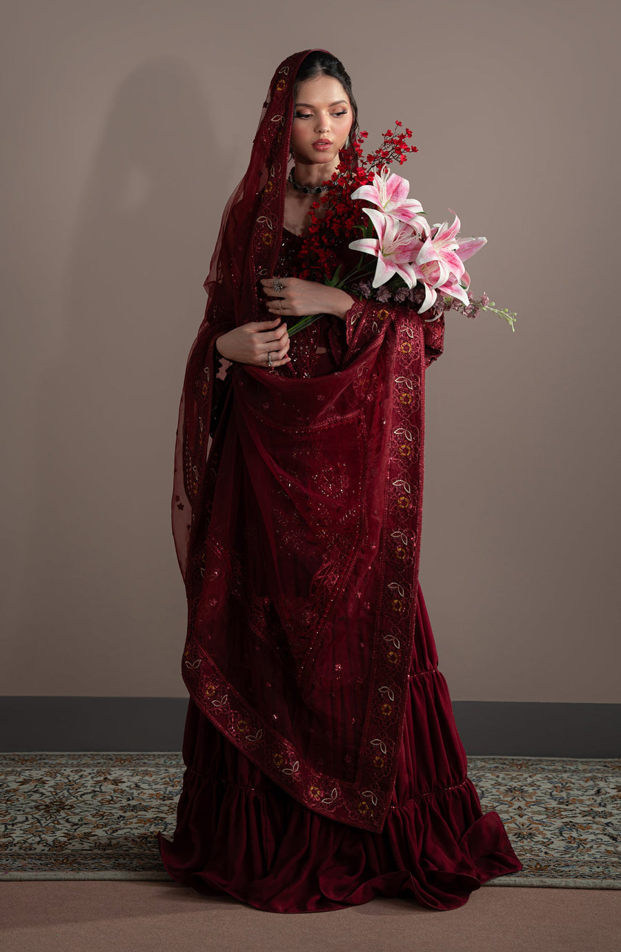 Woman in a burgundy velvet saree holding flowers against a plain background