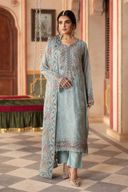 A woman modeling a blue chiffon unstitched 3-piece suit with embroidered details and a shawl, standing in a room with ornate decor.