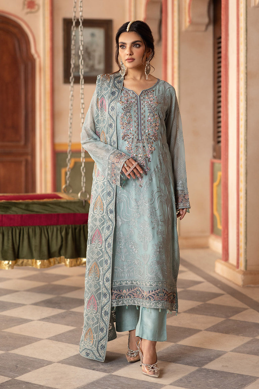 A woman modeling a blue chiffon unstitched 3-piece suit with embroidered details and a shawl, standing in a room with ornate decor.
