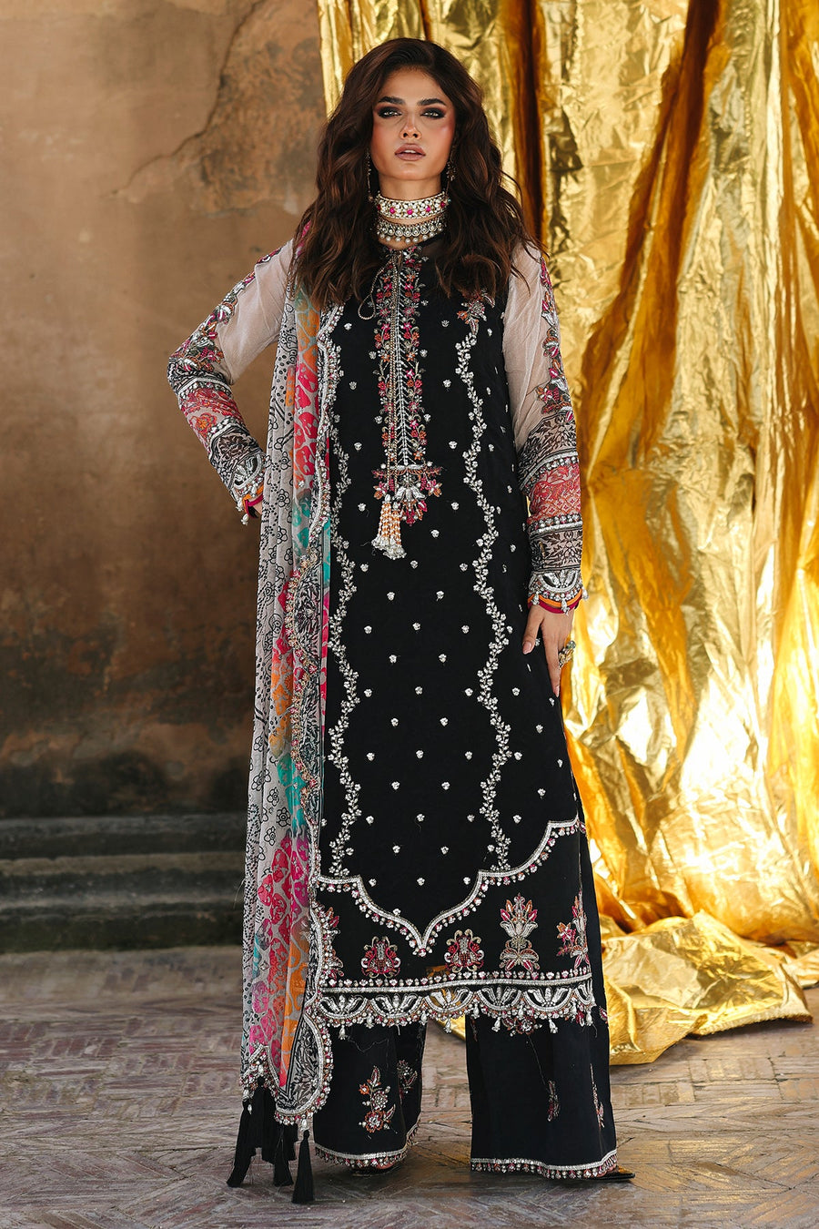 Woman wearing a black embroidered outfit with a colorful scarf against a gold curtain background